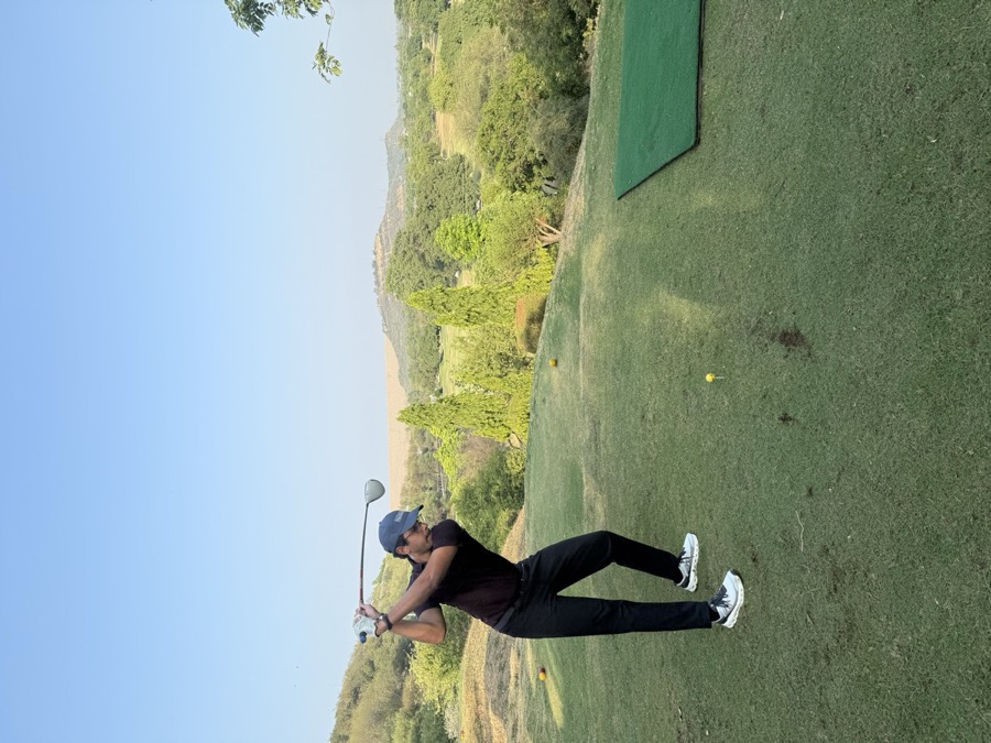 Talha mid swing on a golf tee, hills and trees in the background